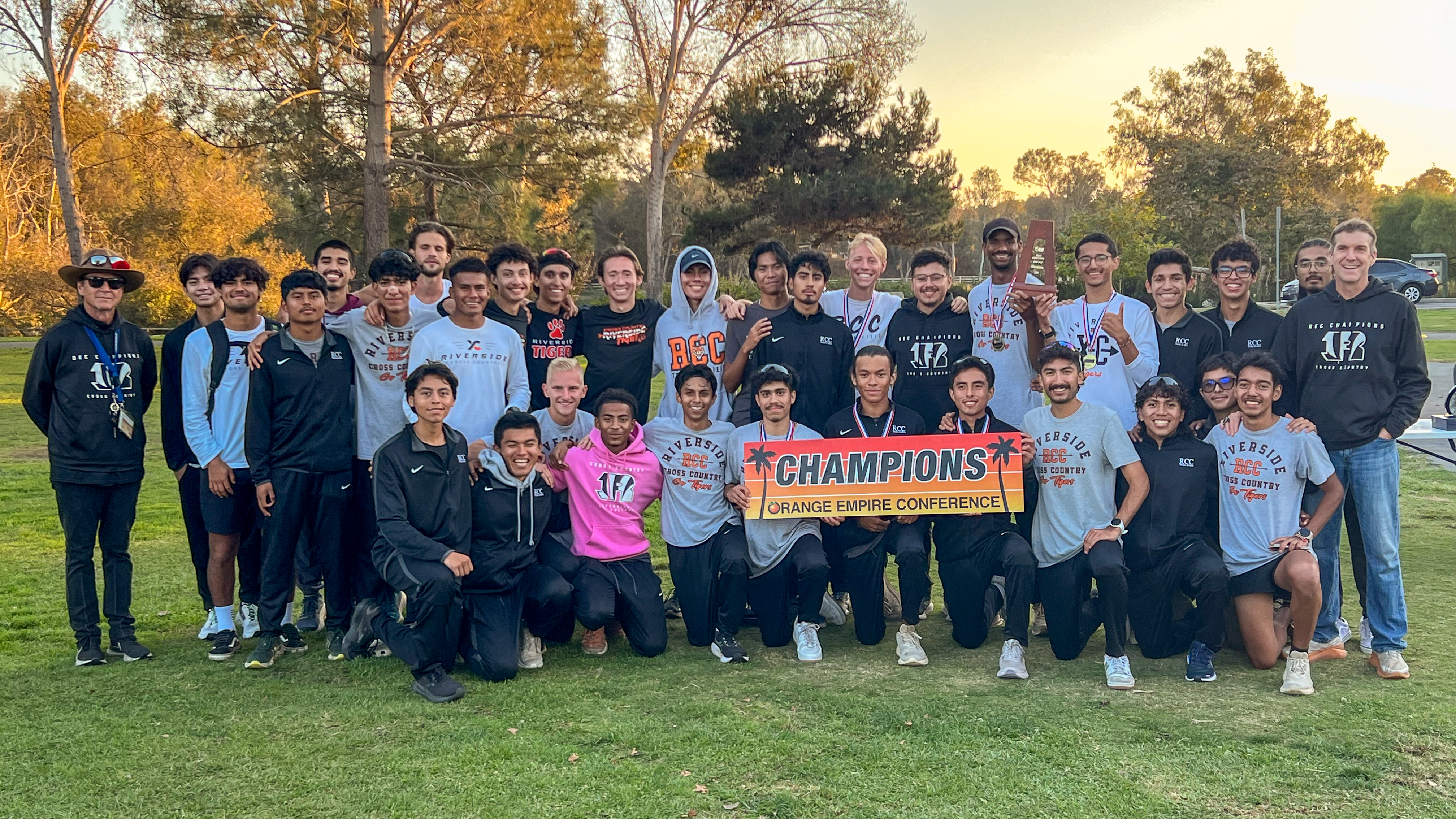 Cross country students holding up a championship banner. Trees and sunset behind them. 