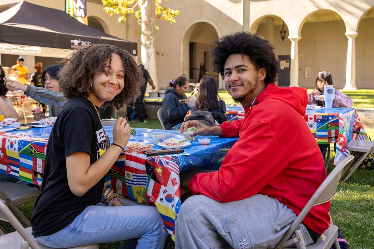 two students eating food from various booths at a table- smiling at the camera