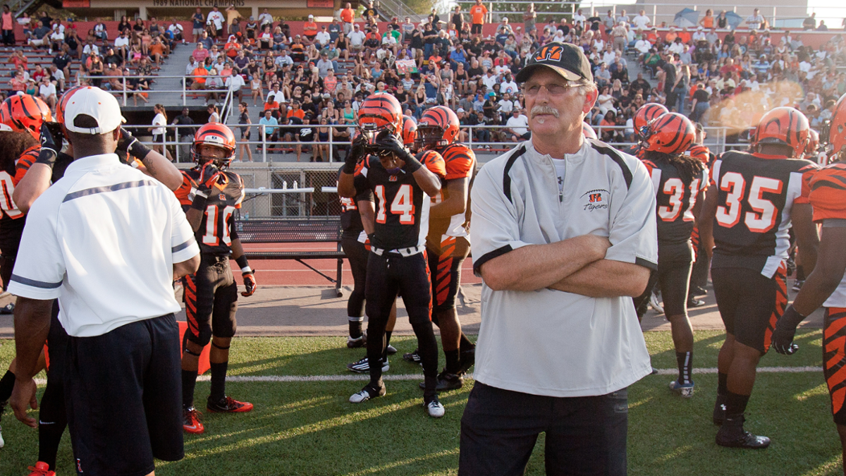 Coach Tom Craft stands with arms crossed on the sideline, looking focused as RCC football players in black and orange uniforms prepare for the game. A crowd of spectators fills the stadium seats in the background.