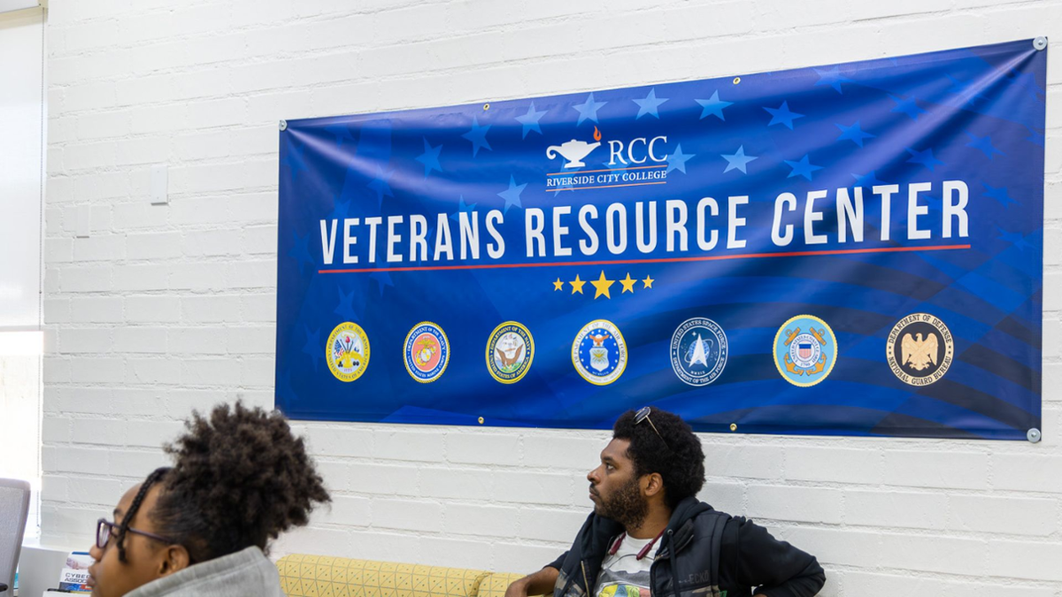 Blue Banner reading Veterans Resources Center against white wall. Two students looking to the left at the bottom