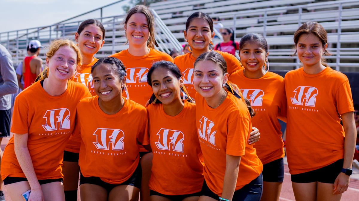 Student smiling with their orange sports shirts in front of bleachers