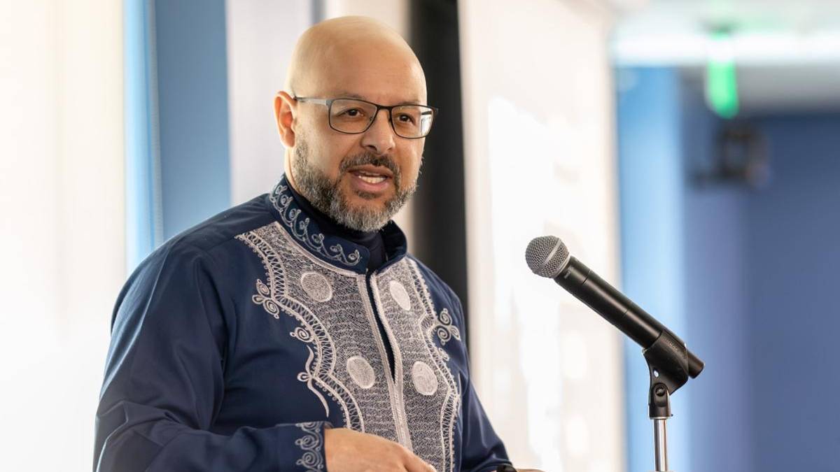 Dr. Nana Chike Akua speaks at a microphone, wearing a traditional blue embroidered shirt, glasses, with a blurred background 