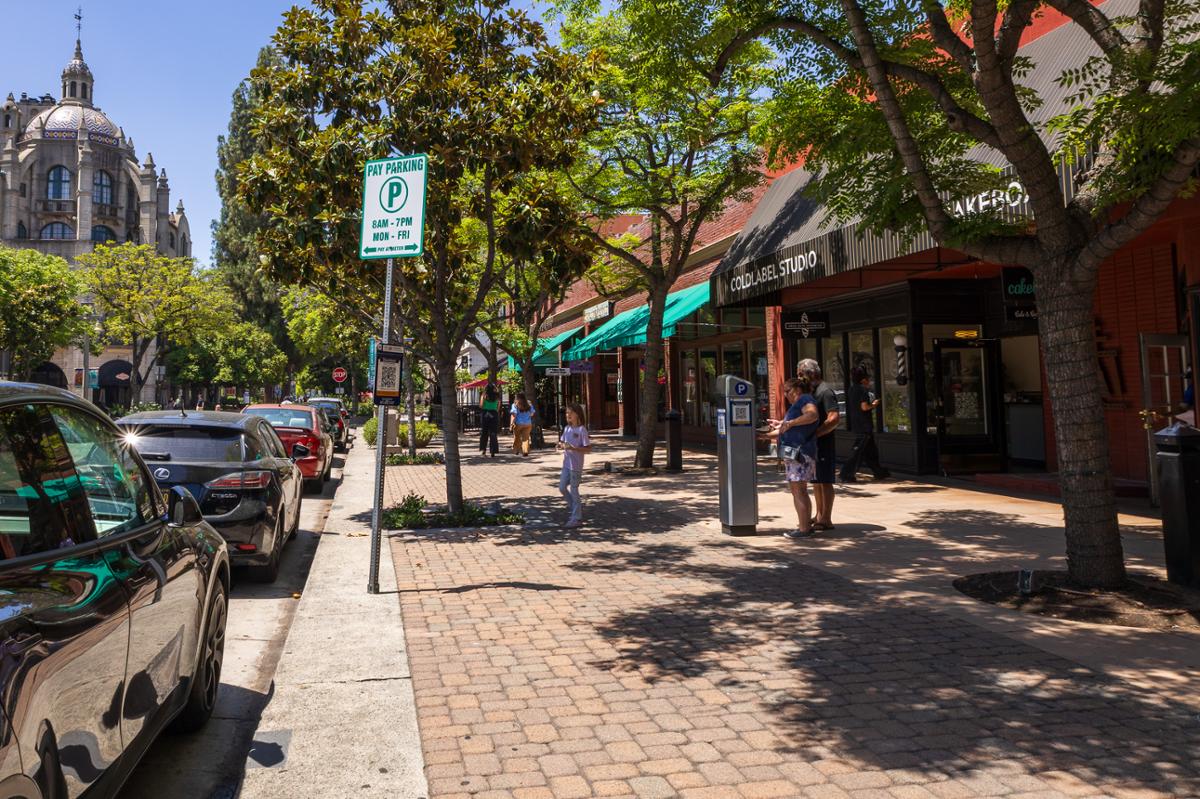 A tree-lined block of downtown Riverside features the storefront of Cold Cutz Barbershop, with pedestrians, parked cars, and nearby businesses along a shaded sidewalk on a sunny afternoon.