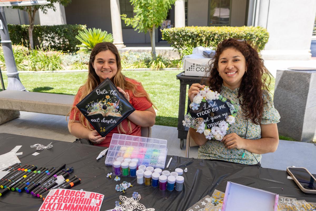 Two RCC students smile while displaying decorated graduation caps at a Grad Fest cap-decorating station. Craft supplies, markers, glitter, and embellishments are spread across the table as they celebrate graduation.
