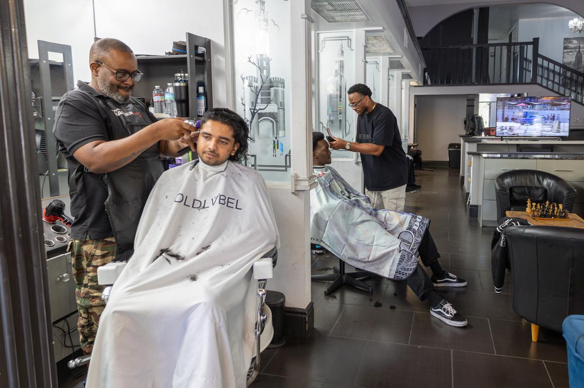 A barber trims a client’s hair while another stylist serves a customer in the background at a local barbershop, highlighting hands-on training and professional grooming services.