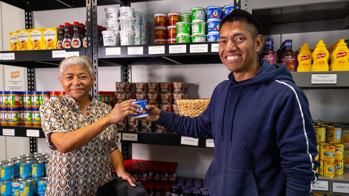 Two people stand inside RCC’s Basic Needs Resource Center pantry, smiling as one hands the other a canned food item. Shelves behind them are stocked with cereal, peanut butter, canned goods, and other grocery essentials for students.
